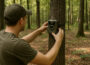 Person zeigt, wie man eine Wildkamera richtig aufstellen sollte, um optimale Aufnahmen im Wald zu erzielen